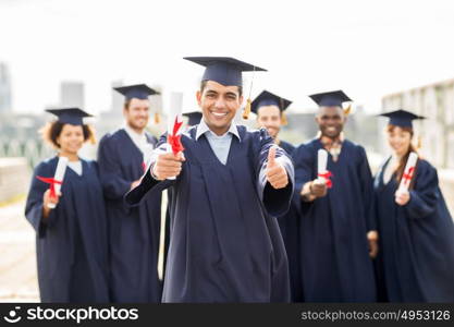 education, graduation, gesture and people concept - group of happy international students in mortar boards and bachelor gowns with diplomas showing thumbs up. happy students with diplomas showing thumbs up