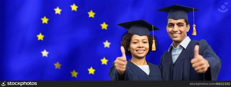 education, graduation, gesture and people concept - happy international graduate students in mortar boards and bachelor gowns outdoors showing thumbs up over flag of european union background. graduate students or bachelors showing thumbs up