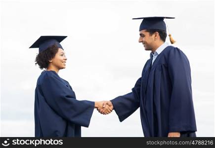 education, graduation, gesture and people concept - happy international students in mortar boards and bachelor gowns greeting each other by handshake outdoors