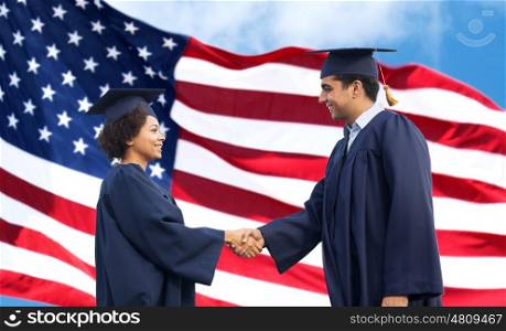 education, graduation, gesture and people concept - happy international students in mortar boards and bachelor gowns greeting each other by handshake over american flag background
