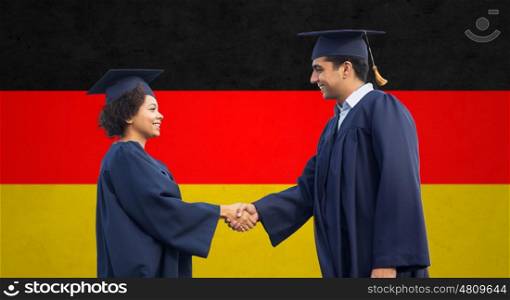 education, graduation, gesture and people concept - happy international students in mortar boards and bachelor gowns greeting each other by handshake over german flag background