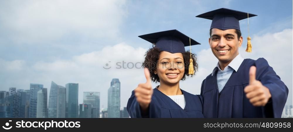 education, graduation, gesture and people concept - happy international students in mortar boards and bachelor gowns outdoors showing thumbs up over singapore city skyscrapers background