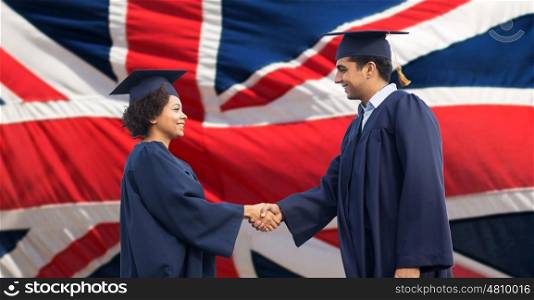 education, graduation, gesture and people concept - happy international students in mortar boards and bachelor gowns greeting each other by handshake over british flag background