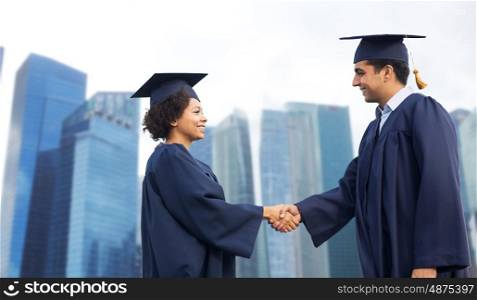 education, graduation, gesture and people concept - happy international students in mortar boards and bachelor gowns greeting each other by handshake over city background