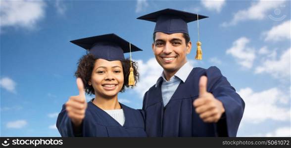 education, graduation, gesture and people concept - happy international students in mortar boards and bachelor gowns outdoors showing thumbs up over blue sky and clouds background. happy students or bachelors showing thumbs up