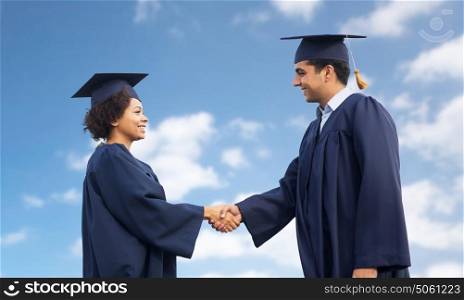 education, graduation, gesture and people concept - happy international students in mortar boards and bachelor gowns greeting each other by handshake over blue sky and clouds background. happy students or bachelors greeting each other