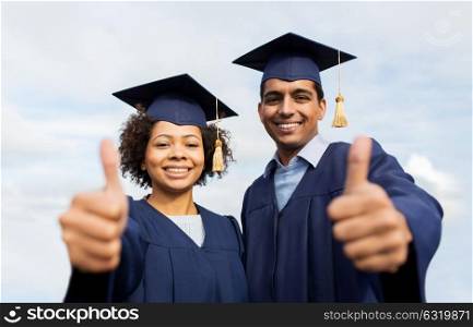 education, graduation, gesture and people concept - happy international students in mortar boards and bachelor gowns outdoors showing thumbs up. happy students or bachelors showing thumbs up