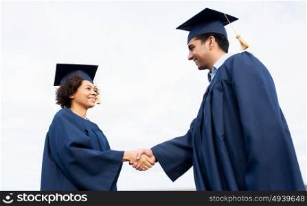 education, graduation, gesture and people concept - happy international students in mortarboards and bachelor gowns greeting each other by handshake outdoors. happy students or bachelors greeting each other