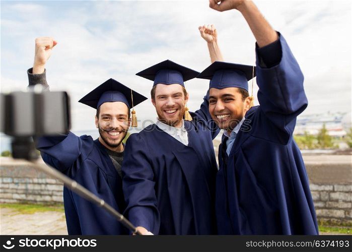 education, graduation, technology and achievement concept - group of happy international students in mortar boards and bachelor gowns taking photo by selfie stick outdoors and celebrating. happy male students or graduates taking selfie