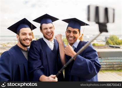 education, graduation, technology and achievement concept - group of happy international students in mortar boards and bachelor gowns taking photo by selfie stick outdoors and celebrating. happy male students or graduates taking selfie. happy male students or graduates taking selfie