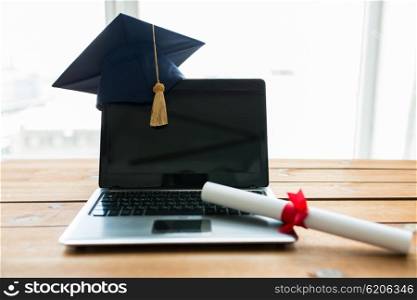education, graduation, technology and e-learning concept - close up of laptop computer with mortarboard and diploma scroll on wooden table