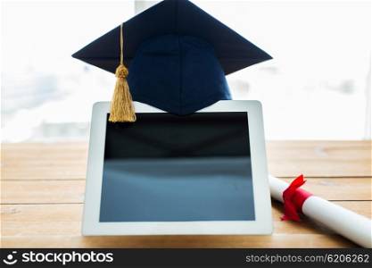 education, graduation, technology and e-learning concept - close up of tablet pc computer with mortarboard and diploma scroll on wooden table