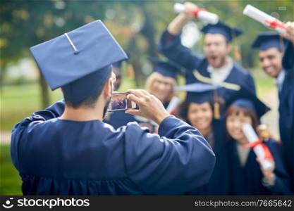 education, graduation, technology and people concept - group of happy international graduate students in mortar boards and bachelor gowns with diplomas taking picture with smartphone outdoors. graduate students taking photo with smartphone