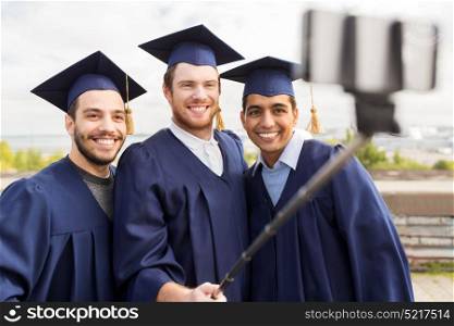 education, graduation, technology and people concept - group of happy international male students in mortar boards and bachelor gowns taking picture by smartphone selfie stick outdoors. happy male students taking picture by selfie stick