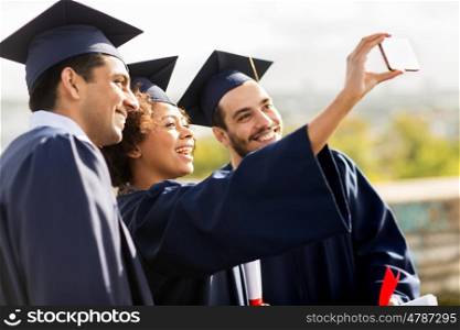 education, graduation, technology and people concept - group of happy international students in mortar boards and bachelor gowns with diplomas taking selfie by smartphone outdoors