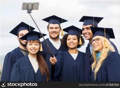 education, graduation, technology and people concept - group of happy international students in mortar boards and bachelor gowns taking selfie by smartphone outdoors. students or bachelors taking selfie by smartphone
