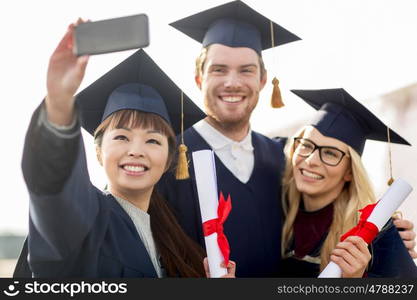 education, graduation, technology and people concept - group of happy international students in mortar boards and bachelor gowns with diplomas taking selfie by smartphone outdoors