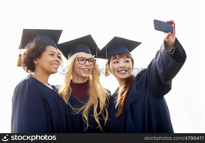 education, graduation, technology and people concept - group of happy international students in mortar boards and bachelor gowns taking selfie by smartphone outdoors