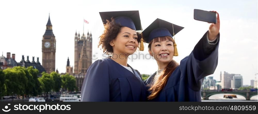 education, graduation, technology and people concept - group of happy international students in mortar boards and bachelor gowns taking selfie by smartphone over london city background