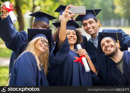 education, graduation, technology and people concept - group of happy international students in mortar boards and bachelor gowns with diplomas taking selfie by smartphone outdoors