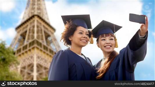 education, graduation, technology and people concept - group of happy international students in mortar boards and bachelor gowns taking selfie by smartphone over eiffel tower background