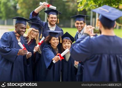 education, graduation, technology and people concept - group of happy international students in mortar boards and bachelor gowns with diplomas taking picture by smartphone outdoors