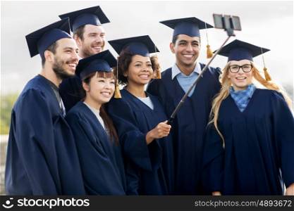 education, graduation, technology and people concept - group of happy international students in mortar boards and bachelor gowns taking selfie by smartphone outdoors