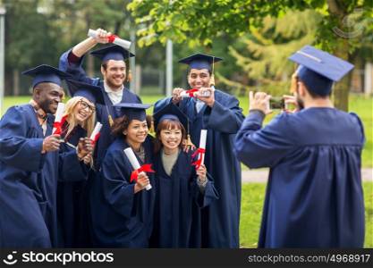 education, graduation, technology and people concept - group of happy international students in mortar boards and bachelor gowns with diplomas taking picture by smartphone outdoors. students or bachelors photographing by smartphone