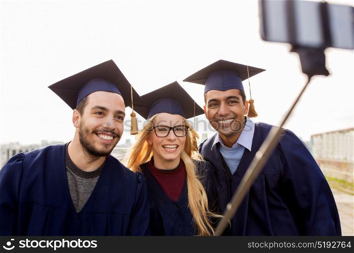 education, graduation, technology and people concept - group of happy international students in mortar boards and bachelor gowns taking selfie by smartphone outdoors. happy students or graduates taking selfie outdoors