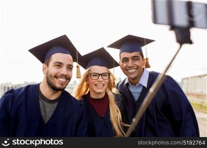 education, graduation, technology and people concept - group of happy international students in mortar boards and bachelor gowns taking selfie by smartphone outdoors. happy students or graduates taking selfie outdoors
