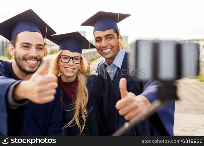 education, graduation, technology and people concept - group of happy international students in mortar boards and bachelor gowns taking selfie by smartphone outdoors and showing thumbs up. students or graduates taking selfie by smartphone