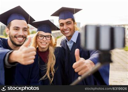 education, graduation, technology and people concept - group of happy international students in mortar boards and bachelor gowns taking selfie by smartphone outdoors and showing thumbs up. students or graduates taking selfie by smartphone