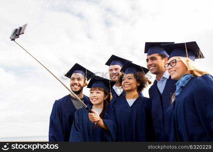education, graduation, technology and people concept - group of happy international students in mortar boards and bachelor gowns taking picture by smartphone selfie stick outdoors. group of happy students or graduates taking selfie