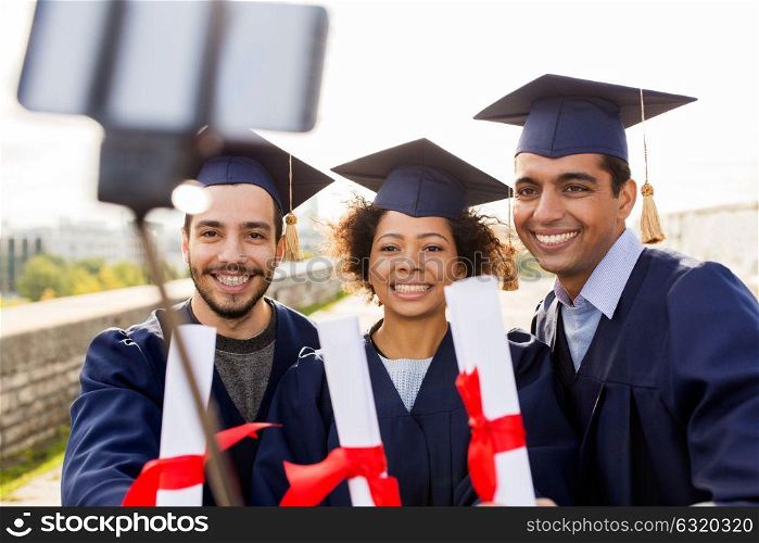 education, graduation, technology and people concept - group of happy international students in mortar boards and bachelor gowns with diplomas taking picture by smartphone selfie stick outdoors. students or graduates with diplomas taking selfie