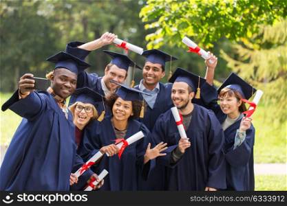 education, graduation, technology and people concept - group of happy international students in mortar boards and bachelor gowns with diplomas taking selfie by smartphone outdoors. students or graduates with diplomas taking selfie
