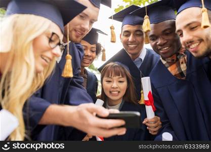 education, graduation, technology and people concept - group of happy international students in mortar boards and bachelor gowns with diplomas and smartphone. students or graduates with diplomas and smartphone