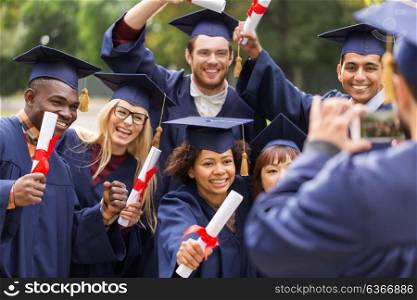 education, graduation, technology and people concept - group of happy international students in mortar boards and bachelor gowns with diplomas taking picture by smartphone outdoors. students or graduates with diplomas taking picture