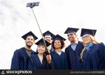 education, graduation, technology and people concept - group of happy international students in mortar boards and bachelor gowns taking picture by smartphone selfie stick outdoors. group of happy students or graduates taking selfie