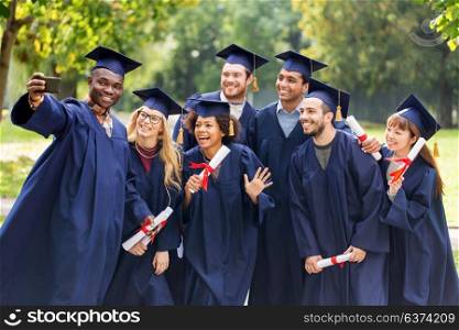 education, graduation, technology and people concept - group of happy international students in mortar boards and bachelor gowns with diplomas taking selfie by smartphone outdoors. students or graduates with diplomas taking selfie