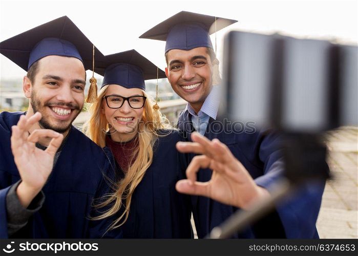 education, graduation, technology and people concept - group of happy international students in mortar boards and bachelor gowns taking selfie by smartphone outdoors and showing ok hand sign. students or graduates taking selfie by smartphone