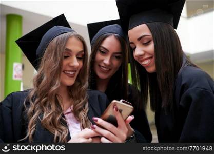 education, graduation, technology and people concept - group of happy international students in mortar boards and bachelor gowns with diplomas using martphone