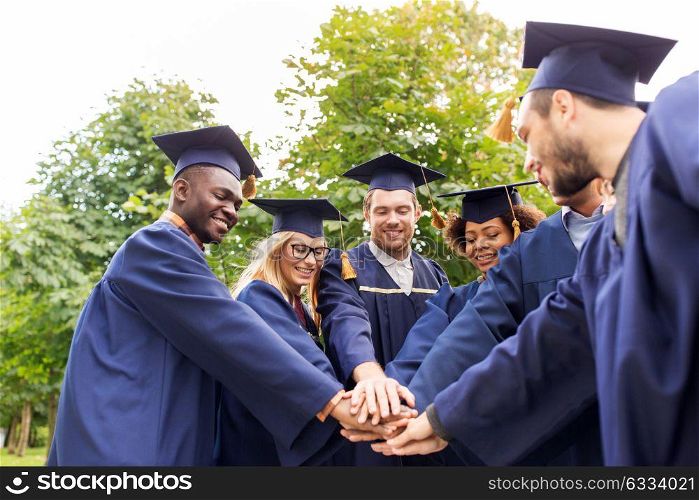 education, graduation, unity and people concept - group of happy international students in mortar boards and bachelor gowns standing in circle and holding hands. happy students or bachelors in mortar boards