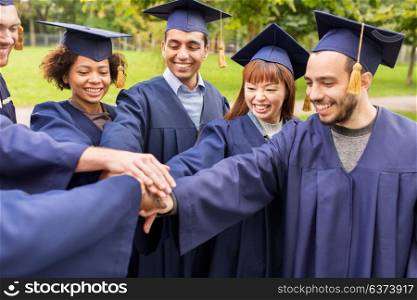 education, graduation, unity and people concept - group of happy international students in mortar boards and bachelor gowns standing in circle and holding hands. happy students or bachelors in mortar boards