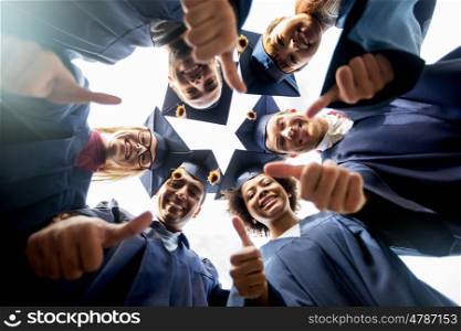 education, graduation, unity, gesture and people concept - group of happy international students in mortar boards and bachelor gowns standing in circle and showing thumbs up