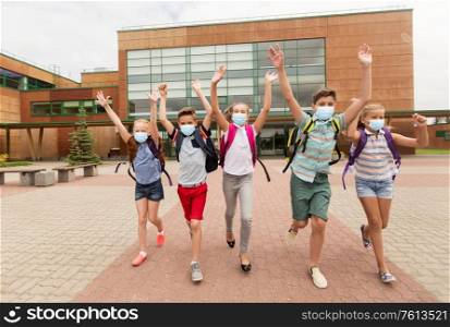 education, healthcare and pandemic concept - group of elementary students wearing face protective medical masks for protection from virus disease with backpacks walking from school and waving hands. group of happy students in masks leaving school