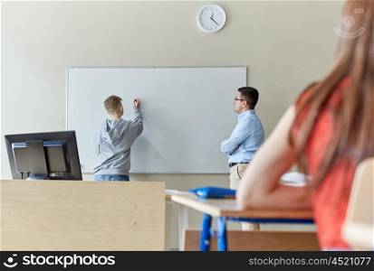 education, high school, learning and people concept - student boy writing something on blank white board and teacher in classroom
