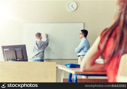 education, high school, learning and people concept - student boy writing something on blank white board and teacher in classroom. teacher and student writing on board at school