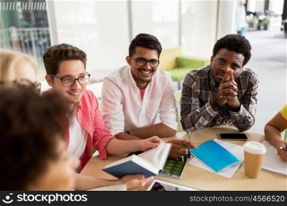 education, high school, people and technology concept - group of international students sitting at table with tablet pc computer, smartphone and notebooks and talking at university