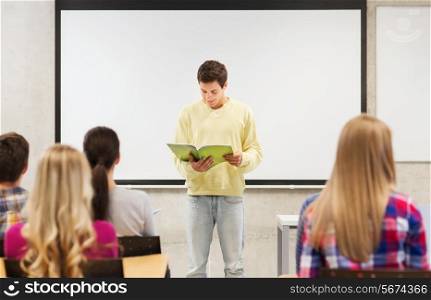 education, high school, teamwork and people concept - smiling student boy with notebook standing and reading in front of students in classroom