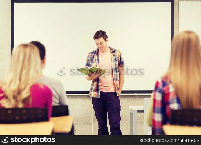 education, high school, teamwork and people concept - smiling student boy with notebook standing and reading in front of students in classroom